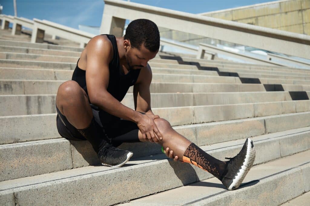 Handsome black runner with muscular athletic body holding his leg with both hands, feeling pain in k