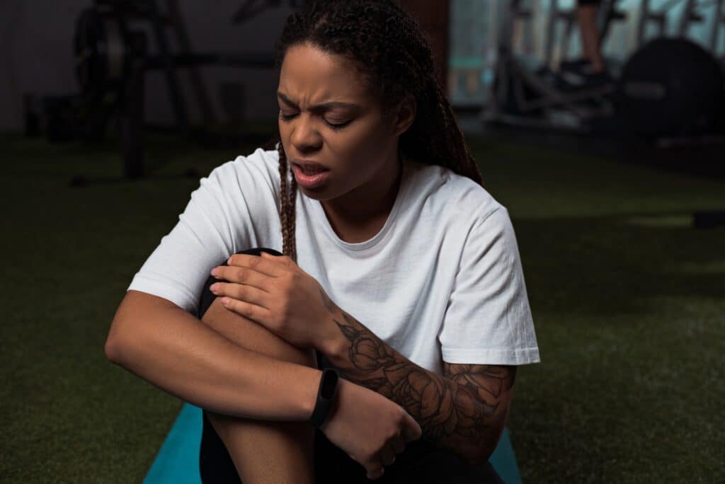 Worried and upset african american woman suffering from pain in knee, sitting on fitness mat
